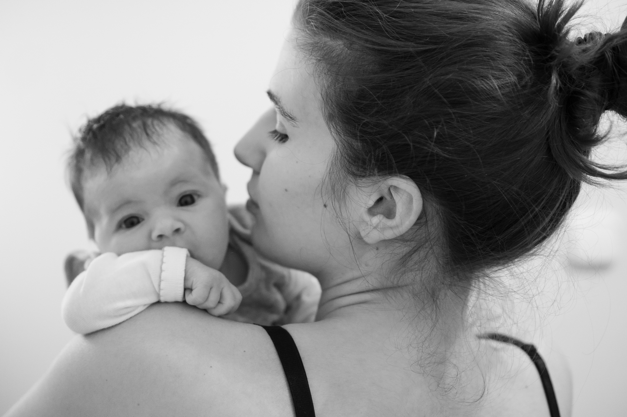 újszülött baba eszi az öklét amíg az anyukája vállán át a kamerába néz